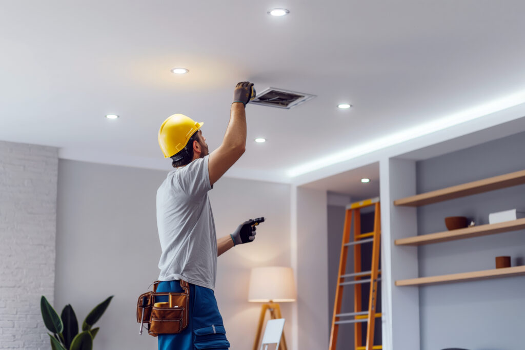 Dans un salon moderne, un électricien professionnel installe un nouveau luminaire au plafond, apportant une touche de style et de fonctionnalité à l’espace. Equipé de sa boîte à outils et de son échelle, il travaille avec précision et expertise pour garantir un résultat impeccable. Cette intervention reflète l'importance de l'entretien et de l'amélioration des installations électriques pour assurer confort et sécurité dans chaque pièce de la maison.